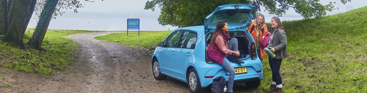 Drie vriendinnen staan om een geparkeerde auto heen. Ze kletsen met elkaar. Ze staan in de natuur met bomen en zand om hen heen.